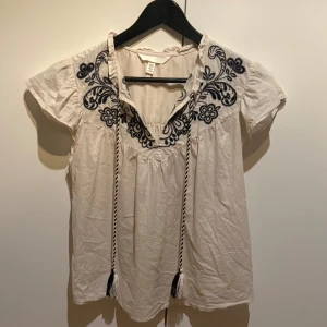 Beige blus med broderi - Supersöt blus från H&M med svart broderat blommönster över axlar och bröst. Superfint skick då den endast är använd vid ett tillfälle😊