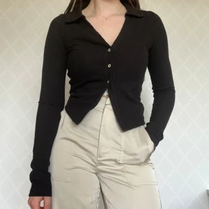 Svart ribbad kofta med krage - Svart ribbad kofta med v-ringning och klassisk krage. Tröjan har lång ärm och knäpps med tre knappar framtill🥰H&M Divided.
