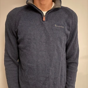 Blå stickad tröja från Bondelid - Mörkblå stickad tröja från Bondelid med halv dragkedja och hög krage. Broderad logga på bröstet och ribbade detaljer vid axlarna. Perfekt för kyliga dagar och enkel att matcha med jeans eller chinos.