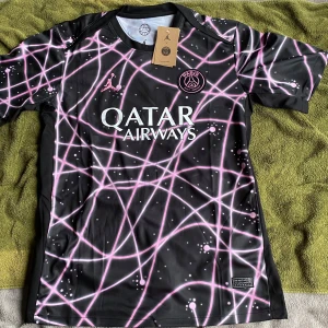 PSG x Jordan svart fotbollströja - Snygg svart PSG fotbollströja i samarbete med Jordan. Tröjan har korta ärmar och är tillverkad i lätt, andasvänligt Dri-FIT-material. Unikt mönster med rosa och vita linjer och prickar över hela tröjan. Qatar Airways-logga på bröstet och PSG-emblem i rosa.