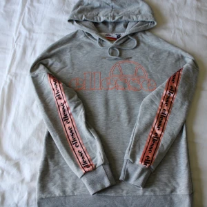 Grå Ellesse hoodie med rosa detaljer - Säljer en grå hoodie från Ellesse med rosa logga framtill och rosa band med Ellesse-tryck längs ärmarna. Tröjan har huva med snörning och ribbade muddar vid ärmslut och nederkant. Perfekt för en chill och sportig look. Buren 2 gånger, väldigt mjuk och gosig. Färgen faller mig inte längre i smak, annars skulle det bli behålla! 