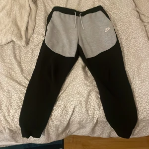 Nike mjukisbyxor svart/grå M - Nike mjukisbyxor i svart med ljusgrå paneler framtill och vit Nike-logga. Byxorna har snörning i midjan, ribbade muddar vid bensluten och är gjorda i mjukt bomullsmaterial. Perfekt för chill eller träning, riktigt skön passform och snygg kontrastdesign.