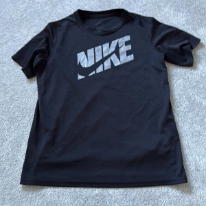 Svart Nike Dri-Fit t-shirt - Svart t-shirt från Nike med stor silverfärgad logga på bröstet. Tillverkad i Dri-Fit-material som andas och håller dig torr. Klassisk passform med rund hals och korta ärmar, perfekt för träning eller chill dagar.