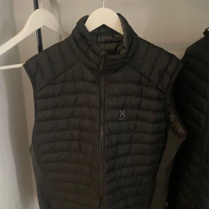 Svart dunväst från Haglöfs - Säljer en svart dunväst från Haglöfs med quiltad design och hög krage. Västen har dragkedja framtill och är perfekt för lager-på-lager. Klassisk och sportig look med diskret logga på bröstet.