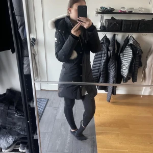 Svart parkas med päls från Moose Knuckles - Säljer en svart parkas från Moose Knuckles med vit päls runt huvan och snygga detaljer på ärmen. Jackan har lång passform, dragkedja och knappar framtill samt fickor. Perfekt för kalla dagar och är väldigt fin.