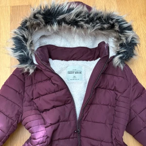 Vinröd pufferjacka från Tally Weijl - Snygg vinröd pufferjacka från Tally Weijl med fuskpäls på luvan och mysigt vitt foder. Jackan har dragkedja framtill, två fickor med dragkedja och quiltade detaljer. Perfekt för kalla dagar när du vill hålla stilen och värmen. Tajt passform med några få skavanker som inte märks till så mycket 💞💞💞