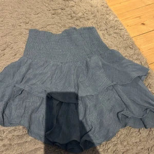 Blå volangkjol med resårmidja - Supersöt blå kortkjol med volanger och bred smockad resår i midjan. Kjolen har en luftig känsla och passar perfekt till varma dagar. Materialet är lätt och ger en flowy look.