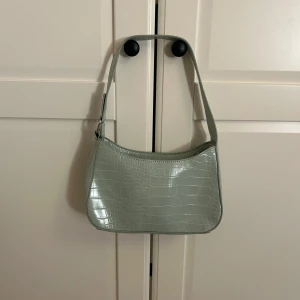 Ljusgrön axelväska i croco-imitation - Snygg ljusgrön axelväska med croco-mönstrad yta och stilren form. Perfekt storlek för det viktigaste och enkel att matcha till olika outfits. Väskan har ett kort axelband och en glansig finish som ger en trendig vibe.