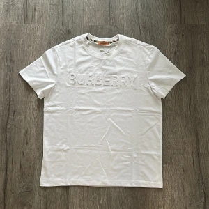 Vit Burberry t-shirt med logotyp - Snygg vit t-shirt från Burberry med stor, upphöjd logotyp på bröstet. Klassisk rund hals och korta ärmar. Tillverkad i mjuk bomull som känns skön mot huden. Perfekt för dig som gillar stilrena designer och vill ha ett statement-plagg i garderoben.