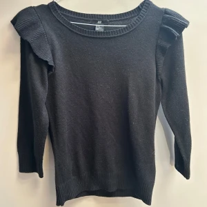 Svart stickad tröja med volang H&M - Svart stickad tröja från H&M med volangdetaljer på axlarna. Tröjan har rund halsringning och trekvartsärm. Perfekt för dig som gillar en enkel men ändå trendig look.