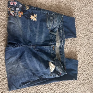 Blå skinny jeans med broderade blommor - Snygga blå skinny jeans från Westport med broderade blommor i olika färger på ena benet. Jeansen har klassisk femficksdesign, knapp och dragkedja fram samt en smal passform som sitter tajt längs benen. Perfekt för dig som vill ha en cool och färgglad detalj på dina jeans.