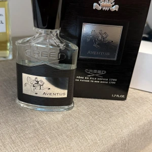 Creed Aventus Eau de Parfum 50ml - Creed Aventus är en lyxig parfym i en elegant, genomskinlig glasflaska med svart och silverdetaljer. Flaskan har en svart etikett med en ryttare och texten 'Aventus'. Parfymen är 50 ml och har en stilren svart kork. Perfekt för dig som gillar exklusiva dofter.