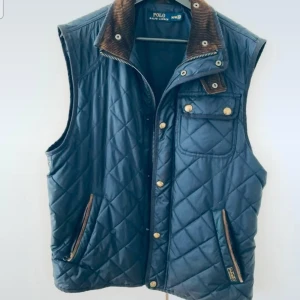 Quiltad blå dunväst från Polo Ralph Lauren - Snygg quiltad blå dunväst från Polo Ralph Lauren med bruna detaljer i manchester på kragen. Västen har dragkedja, tryckknappar, fickor med dragkedja och en bröstficka med lock. Perfekt lager-på-lager-plagg för kyligare dagar.