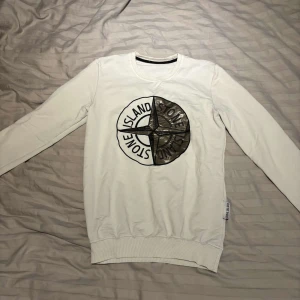 Vit långärmad tröja från Stone Island - Vit långärmad tröja från Stone Island med stort svart och grått logotryck på bröstet. Tröjan har rund halsringning, ribbade muddar och är tillverkad i mjuk bomull. Perfekt för dig som gillar streetwear och vill sticka ut med en ikonisk look.