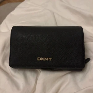 Svart plånbok från DKNY i skinn - Snygg svart plånbok från DKNY i strukturerat skinn med guldfärgad logga framtill. Plånboken har flera fack för kort, mynt och sedlar samt två dragkedjefack och ett fönster för ID. Perfekt för att hålla ordning på allt viktigt.