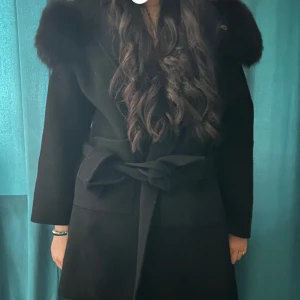 Svart kappa med pälsdetalj och bälte - Snygg svart kappa med bred pälsdetalj runt kragen och ett knytbälte i midjan. Jackan har en klassisk, rak form och långa ärmar. Perfekt för dig som vill ha en elegant och varm look under höst och vinter. (Äkta päls). Priset kan dessutom diskuteras.