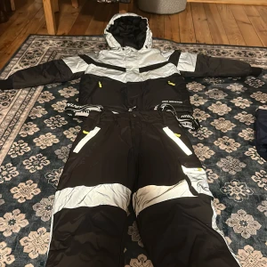 Svarta och grå täckbyxor och vinter jacka High Mountain - Säljer ett par svarta och grå täckbyxor och liknande vinterjacka från High Mountain med justerbara hängslen och flera fickor med dragkedja och knapp. Byxorna har breda gråa partier och detaljer i gult. Perfekta för vinterns alla äventyr i snö och kyla.