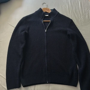 Mörkblå stickad kofta från DeFacto - Säljer en mörkblå stickad kofta från DeFacto i storlek XS. Koftan har grovstickad struktur, hel dragkedja framtill och ribbade muddar vid ärmslut, nederkant och krage. Perfekt för lager-på-lager under kyliga dagar. Pass form som S