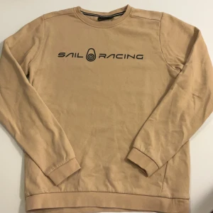 Beige Sail Racing sweatshirt - Snygg original beige sweatshirt från Sail Racing med svart logotyp tryckt på bröstet och en mindre logga i nacken. Tröjan har rund hals, ribbade muddar och är tillverkad i mjuk bomull. Perfekt för chill dagar och enkel att matcha med jeans eller joggers. Storlek 170 (passar strl S). Varans namn: JR bowman sweater, nypris: 6-700kr 
