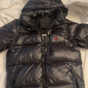 Svart dunjacka från Ralph Lauren - Snygg svart dunjacka från Ralph Lauren med glansig finish och röd logga på bröstet. Jackan har huva, dragkedjor framtill och på fickorna samt quiltad design. Perfekt för kalla dagar och riktigt stilren look.