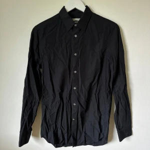 Svart slim fit skjorta från Arket - Svart skjorta från Arket i slim fit-modell, tillverkad i tunn och mjuk bomullspoplin. Klassisk krage, långa ärmar med manschett och knäppning framtill. Enkel och stilren design som passar perfekt till jeans eller kostymbyxor. 1F6