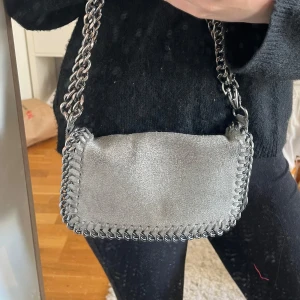 Silver axelväska med kedjedetaljer - Snygg axelväska i silverfärgad mocka med grova kedjor som axelrem och runt kanterna. Väskan har en mjuk, rektangulär form och ger en cool vibe till din outfit. Perfekt storlek för det viktigaste och riktigt trendig detalj.