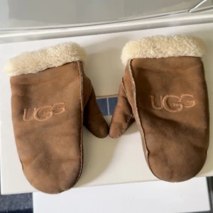 Bruna UGG tumvantar i mocka - Säljer ett par bruna tumvantar från UGG med mjukt vitt foder och broderad logga på ovansidan. Vantarna är i mocka och har en klassisk, enkel design med uppvikt kant i fårskinn. Perfekta för kalla vinterdagar.
