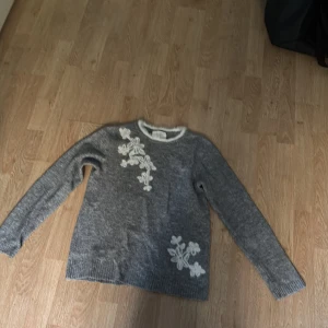 Grå stickad tröja med blommor Springfield - Grå stickad tröja från Springfield i storlek S. Tröjan har vita broderade blommor på framsidan och en vit kant vid halsen. Långärmad och mjuk, perfekt för kyliga dagar. Enkel och snygg design som passar till jeans eller kjol.