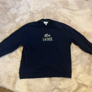Marinblå sweatshirt från Lacoste - Säljer en stilren marinblå sweatshirt från Lacoste med klassisk krokodillogo och vit text på bröstet. Tröjan har rund hals, långa ärmar och mjukt material som är skönt mot huden. Perfekt för dig som gillar enkel och snygg streetstyle.
