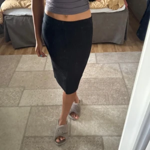 Svart ribbad stickad långkjol - Säljer en svart, ribbad stickad långkjol med slim passform. Kjolen är midilång och sitter snyggt runt kroppen. Perfekt att styla med en croppad topp eller fin blus beroende på tillfälle. Materialet är mjukt och stretchigt, vilket gör den superbekväm att bära hela dagen.