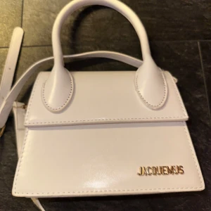Vit Jacquemus handväska i skinn - Superfin vit handväska från Jacquemus i slät skinn med guldiga metalldetaljer och logga framtill. Klassisk miniform med topphandtag och avtagbar axelrem. Perfekt för dig som gillar stilrena accessoarer med lyxig känsla.