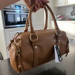 Beige handväska i veganskt material - Snygg beige handväska i veganskt material med struktur och två rejäla handtag. Väskan har guldfärgade metalldetaljer, dragkedja upptill och på sidorna samt en stilren, rundad form. Perfekt för dig som vill ha en trendig och praktisk väska.