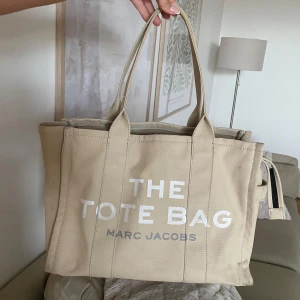 Beige shoppingväska Marc Jacobs - Säljer en beige Tote bag från Marc Jacobs i canvas med tryckt text på framsidan. Väskan har två långa handtag, dragkedja upptill och rymligt innerfack. Perfekt för dig som vill ha en stilren och trendig tote bag med plats för allt du behöver.