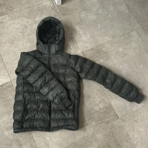 Mörkgrön dunjacka Peak Performance 170 - Säljer en svart dunjacka från Peak Performance med huva och dragkedja. Jackan är quiltad och har två fickor med dragkedja. Perfekt för kalla dagar och har en clean, sportig look. Passar dig som gillar stilren och funktionell design.