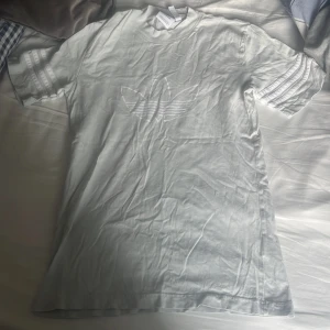  Adidas t-shirt  - Säljer en vit t-shirt från Adidas med det klassiska trefoil-loggan ton-i-ton på bröstet och tre ränder på ärmarna. T-shirten är i mjuk bomull och har en avslappnad passform. Perfekt för dig som gillar sportig och clean stil.
