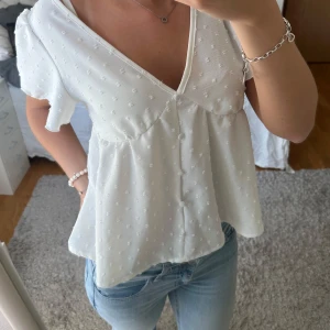 Vit blus med v-ringning och knappar - Supersöt vit blus med v-ringning och korta ärmar. Blusen har små broderade prickar i tyget, knappar framtill och en luftig, flowy passform. Perfekt till jeans eller kjol för en fräsch look. Materialet känns lätt och somrigt.