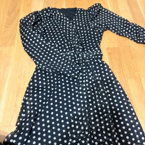Svart prickig långklänning med knyt - Snygg svart långklänning med vita prickar och knyt i midjan. Klänningen har långärmade puffärmar och v-ringad hals. Perfekt för dig som gillar retrovibbar och vill sticka ut med ett coolt mönster. Materialet känns lätt och luftigt.