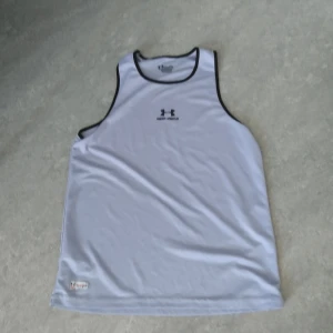 Vit linnetopp från Under Armour XL - Sportig vit linnetopp från Under Armour med svarta kanter och logga framtill. Perfekt för träning eller varma dagar, tillverkad i lätt och snabbtorkande polyester. Klassisk ärmlös design med rund halsringning.