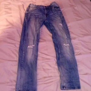 Blå slitna jeans med raka ben - Säljer ett par blå jeans med slitningar och hål på benen för en cool, avslappnad look. Jeansen har klassisk femficksdesign och raka ben. Perfekta för dig som gillar en streetig stil.