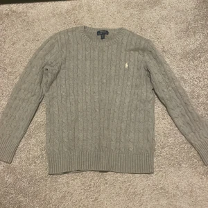 Grå kabelstickad tröja Polo Ralph Lauren - Säljer en grå kabelstickad tröja från Polo Ralph Lauren med klassisk rund hals och långärmad passform. Tröjan har det ikoniska broderade Polo-logot på bröstet och ribbade muddar vid ärmslut och nederkant. Perfekt för dig som gillar stilren och tidlös design. Skick skulle jag säga 9/10. Storlek är 14-16 år vilket är L i barnstorlek