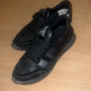 Valentino Garavani svarta sneakers - Svarta Valentino Garavani sneakers med diskret camouflage-mönster. Skorna har snörning, rund tå och platt sula. Materialmix av skinn och mocka ger en lyxig känsla. Perfekta för dig som gillar streetstyle med exklusiv touch.