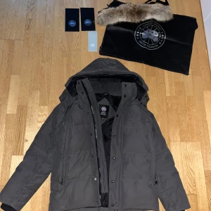 Grå Canada Goose Jacka - Säljer en mörkgrå dunjacka från Canada Goose med avtagbar pälskrage och huva. Jackan har klassisk patch på ärmen, dragkedja och knappar framtill samt två sidofickor. Perfekt för kalla vinterdagar och riktigt snygg streetstil.