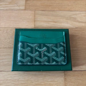 Goy4rd Plånbok - Säljer en grön plånbok med deras ikoniska grafiska mönster i vitt och svart. Plånboken har flera kortfack och är tillverkad i skinn med en lyxig känsla. Perfekt för dig som vill ha något unikt och stilrent i fickan.