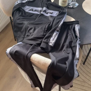 Askari tracksuit limited edition - Svarta mjukisbyxor från Askarpi med breda gråa sidopaneler och logga längs benen. Byxorna har resår i midjan och dragkedjefickor. Perfekta för chill eller träning, med sportig vibe och bekväm passform.
