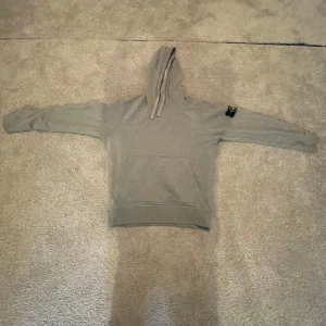 Grå hoodie från Stone Island M - Snygg grå hoodie från Stone Island med klassisk patch på vänster ärm. Tröjan har huva med snörning och är tillverkad i mjuk bomull. Perfekt för dig som gillar streetwear och vill ha en stilren look.