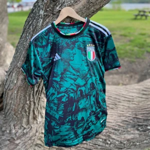 Italien fotbollströja Adidas  - Snygg Italien fotbollströja från Adidas i slim fit, storlek S. Tröjan har ett unikt konstnärligt mönster med motiv från italiensk kultur i mörkgrön och svart, samt vita Adidas-ränder på axlarna. Rund hals med detaljer i rött, vitt och grönt. Perfekt för dig som gillar stil och fotboll.