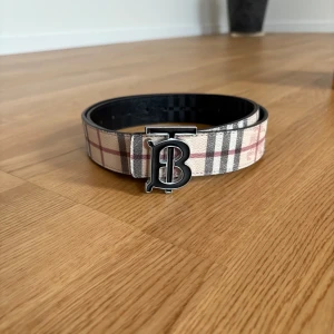Burberry bälte - Snyggt Burberry-bälte med klassiskt rutigt mönster i beige, svart, rött och vitt. Bältet har ett stort metallspänne med Burberry B-logga. Tillverkat i skinn och levereras med originalförpackning, dustbag och äkthetskort. (Förpackning ingår inte)