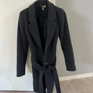Mörkgrå kappa med bälte från H&M - Mörkgrå kappa från H&M med krage, knappar och knytbälte i midjan. Storlek 34