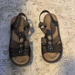 Bruna sandaler från Rieker med pärldetaljer - Snygga bruna sandaler från Rieker med elastiska band och dekorativa pärlor i svart, vitt och silver på ovansidan. Skorna har platt sula och öppen tå, perfekt för varma dagar. Bekväma och stilrena med coola detaljer som sticker ut.