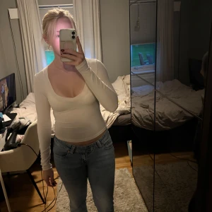 Vit långärmad topp - Säljer en stilren vit långärmad topp med djup rundringning. Toppen är figurnära och tillverkad i ett stretchigt, tunt material som sitter snyggt på kroppen som är mot flishållet. Perfekt att matcha med jeans eller kjol.